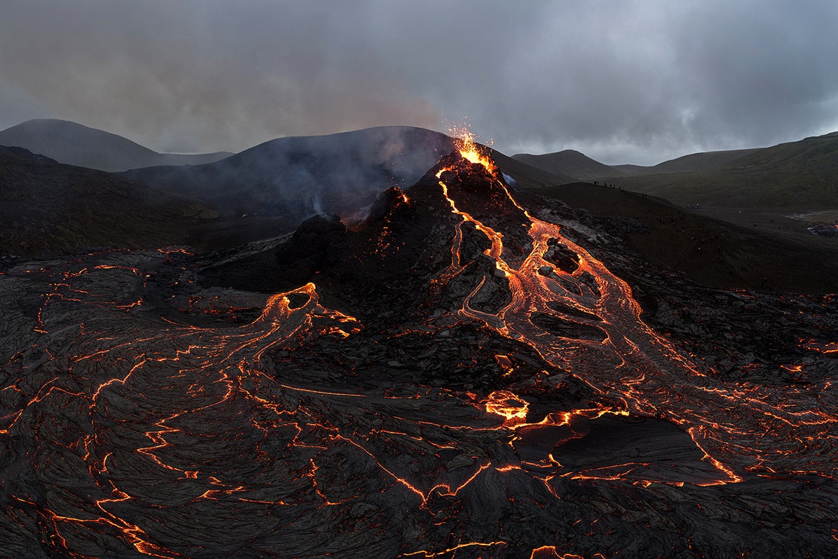 Dark and moody landscape with bright yellow-red lava flowing out of and down the side of a volcano.