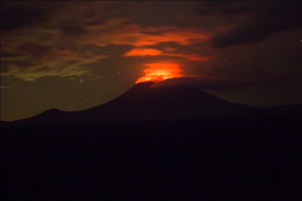 Image shows a volcano in silhouette, rearing against the sky. An orange glow from the summit reflects from the clouds. There is a scattering of stars in the clear bits of sky.