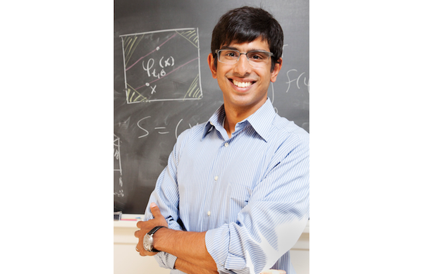 Mathematician Jayadev Athreya is smiling at the camera. He is Indian American, has black hair, and is wearing glasses and a light blue button-front shirt. He is standing in front of a blackboard with math symbols and drawings on it.