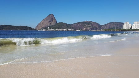 A wave crashes on a beach. Beyond the water, a large black rock formation comes out of the water.