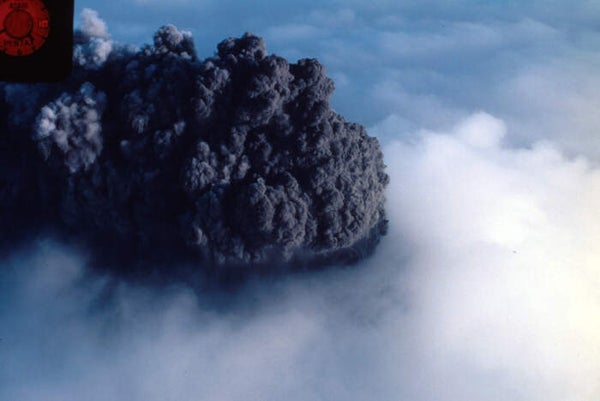 Aerial image shows a gray-black plume of ash boiling above the white cloud ceiling.