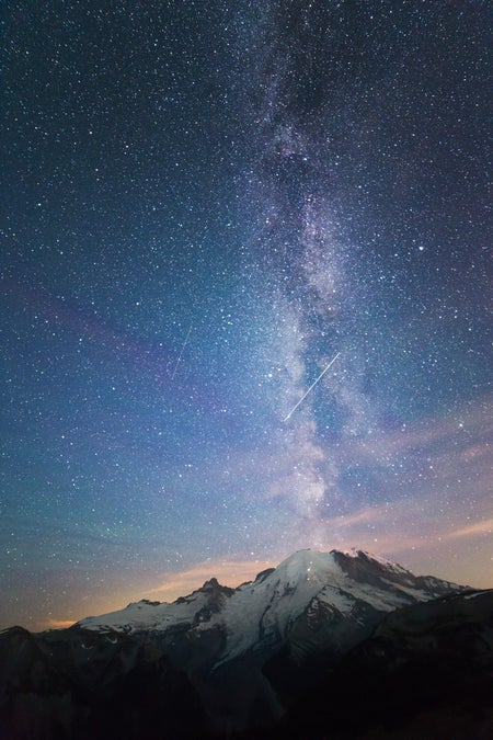 Image shows Mount Rainier with the Milky Way rising over it in a blue and pink sky. There are streaks from meteorites along with the riot of stars. Little Tahoma peak is also visible..