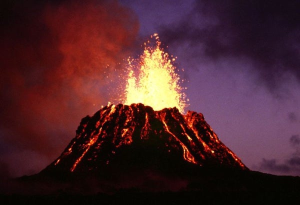 Image shows an erupting volcanic cone at night. It has a flat top, with red lava streaming down the sides, and orange lava bursting from the top.