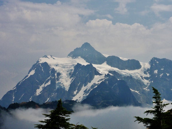 Image shows the jagged summit of Mount Baker, covered in snow and surrounded by clouds.