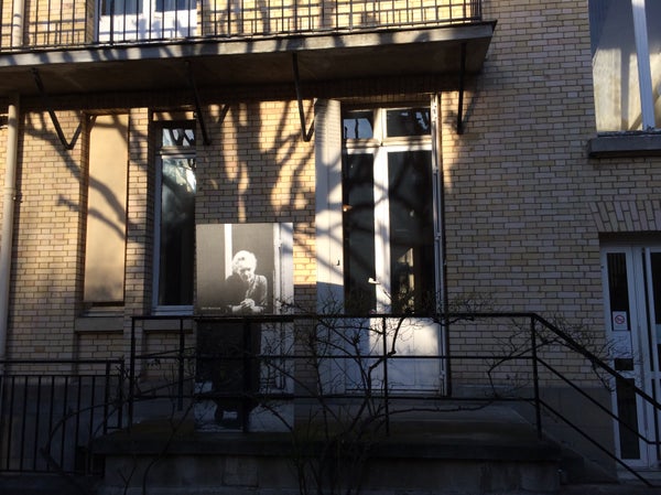 Image description: A life-size photograph of Marie Curie leans against a metal rail outside a brick building.