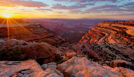 On the left, the sun sets on red rock mesas, sending streaks of red-gold light over them. The rest of the photo looks from a juniper-studded mesa down to canyons and pinnacles.