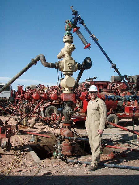 Image shows a man in tan overalls and a white hard hat standing beside a large fracking well head, which basically looks like a Rube Goldberg device made from fire hydrants, pipes, and metal wheels, piled atop each other and sticking out of a hole in the