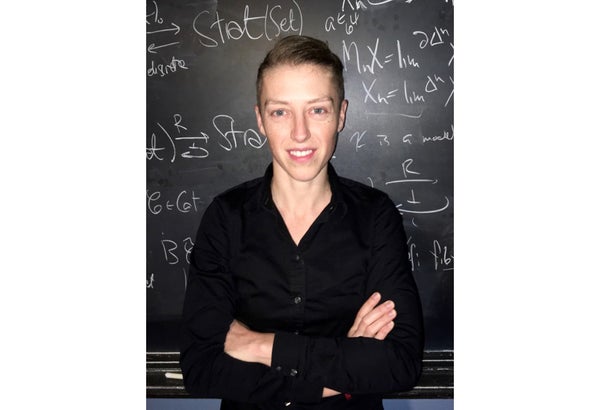 Emily Riehl stands in front of a blackboard filled with mathematics. She is a white woman with short blonde hair wearing a black button-front shirt.