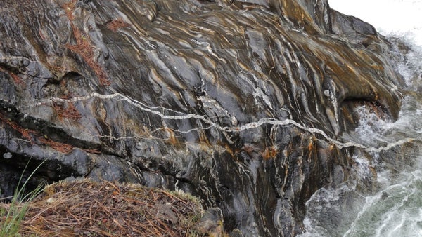 Image shows a wet black schist slope with a white quartz vein running through it.