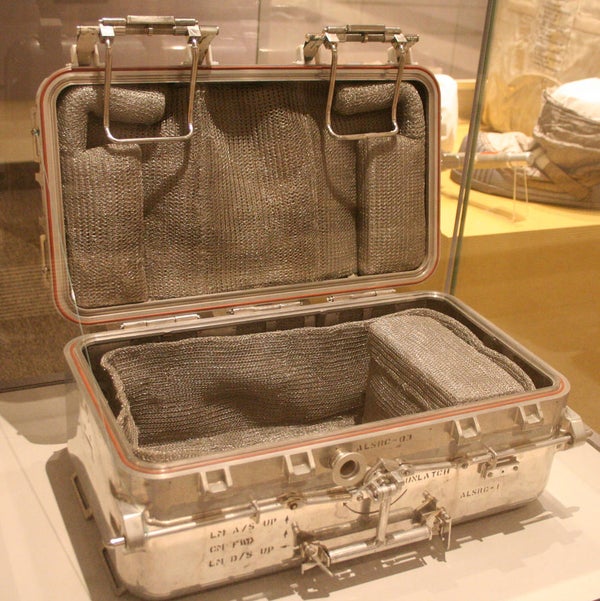 Image shows an open metal case that resembled a small, fat suitcase. It's lined with a knitted mesh. The interior has a large open space and a smaller rectangular case covered in the same mesh on the right side.