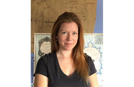 A woman with long red hair wearing a black t-shirt stands in front of posters showing the Mandelbrot set.