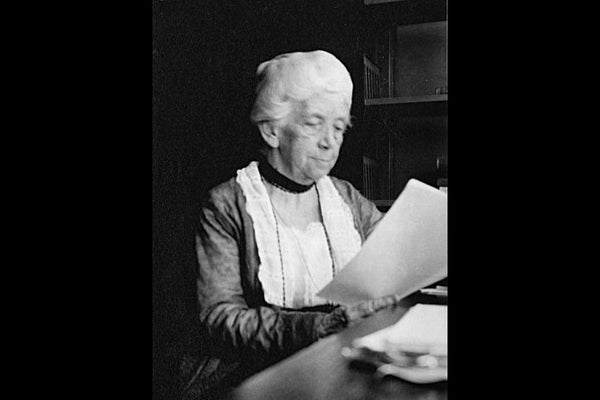 A black and white photograph of a white woman, probably in her 60s or 70s, sitting at a desk reading from a piece of paper