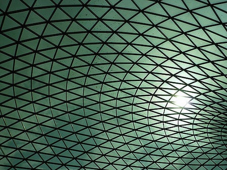 A view up at the roof of the British museum. The sky looks greenish blue behind a black triangular grid. The sun is causing a glare in the right side of the picture.