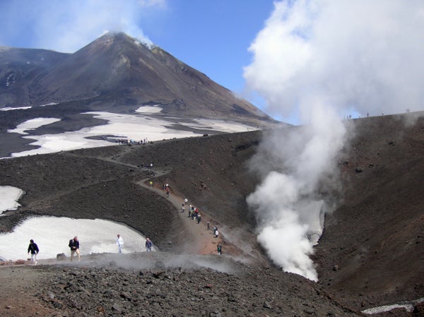 Image shows the steaming summit of Mount Etna in the background. In the foreground, a trail winds between snowfields and craters. Both the summit and the crater in the near right of the photo are steaming. The landscape is nothing but volcanic rock and as