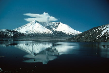 Image shows a snow-shrouded Mount St. Helens reflected in the calm waters of Spirit Lake. A plume of steam rises from the center of her crater: the top of it has been blown by the wind into anvil-shaped clouds close to the summit.