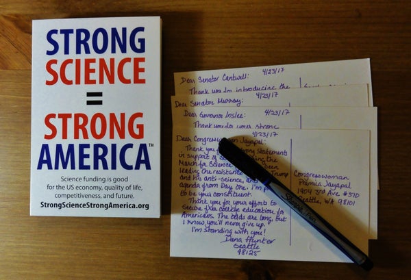 Image shows a stack of Strong Science = Strong America postcards on a wooden surface. Beside them is a set of postcards written to various politicians in purple ink. The Sharpie pen is lying across them.