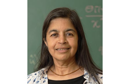 A shot of Nalini Joshi from the shoulders up. She is looking at the camera and smiling. She has long black hair, brown eyes, and brown skin. She is wearing a small brown beaded necklace and a white and blue button-front shirt over a black shirt.
