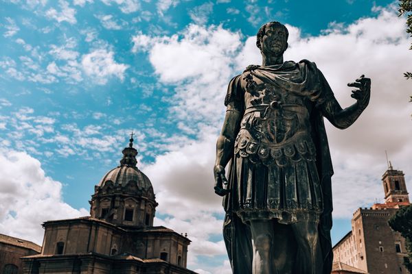 Large bronze statue in the foreground is of Julius Caesar with a domed building in the background beneath a bright blue sky with puffy clouds.
