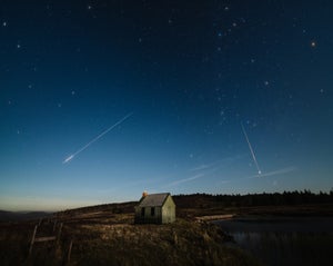 Several meteors streak across the night sky over a small house in the countryside