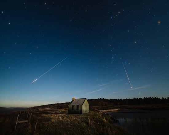 Several meteors streak across the night sky over a small house in the countryside