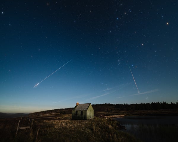 Several meteors streak across the night sky over a small house in the countryside