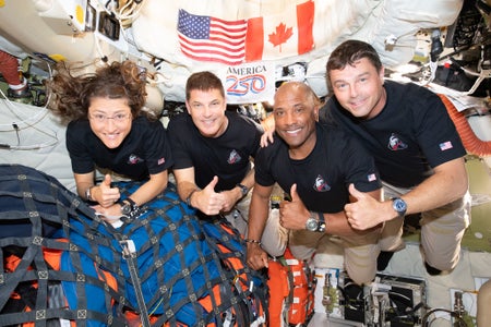 Four people make thumbs-up signs inside a tight spacecraft.