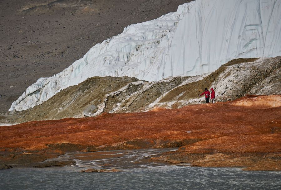 People stand on the bright orange-red ground of Blood Falls, with brown, snow- and ice-covered landscape in the background