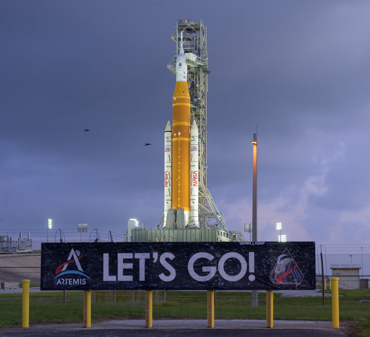 A Space Launch System rocket and Orion crew capsule sit on a pad, awaiting launch. A banner with the words “LET’S GO!” appears in the foreground.