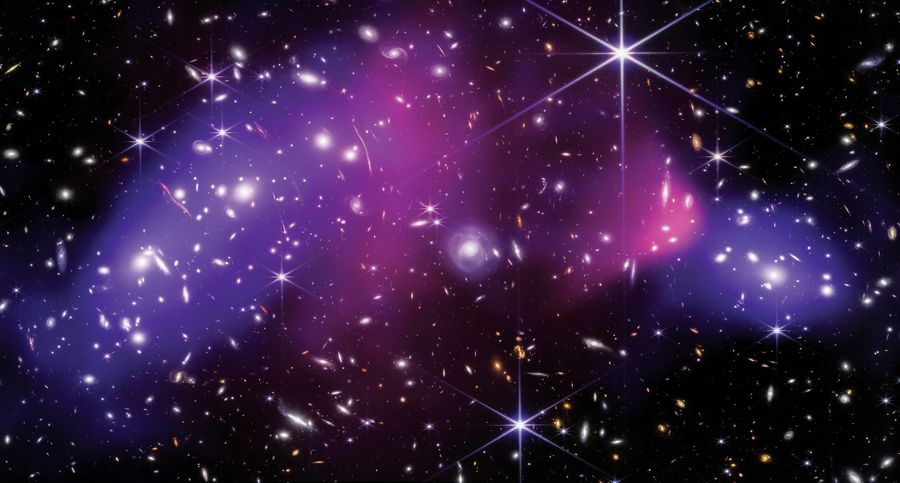 Against a field of distant galaxies, two pink clouds at the center of the image represent regular matter, while a blue cloud on either side represents the unseeable dark matter of the Bullet Cluster. Provided by NASA, ESA, CSA, STScI, CXC; Science: James Jee/Yonsei University, U.C. Davis, Sangjun Cha/Yonsei University, Kyle Finner/Caltech/IPAC