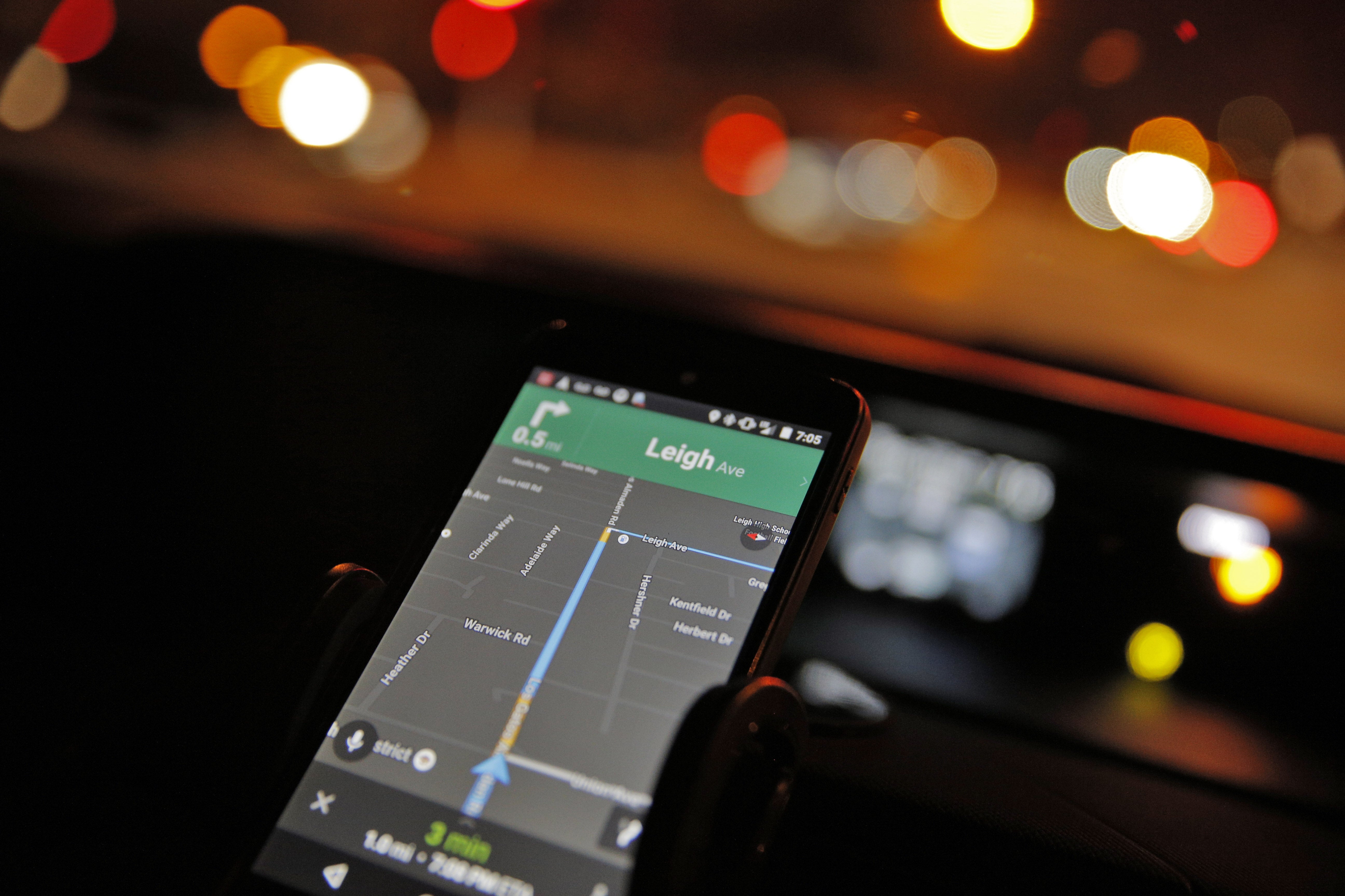 A smartphone mounted in a car displays a navigation map as blurred lights appear through the windshield at night.