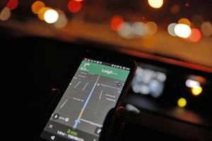 A smartphone mounted in a car displays a navigation map as blurred lights appear through the windshield at night.