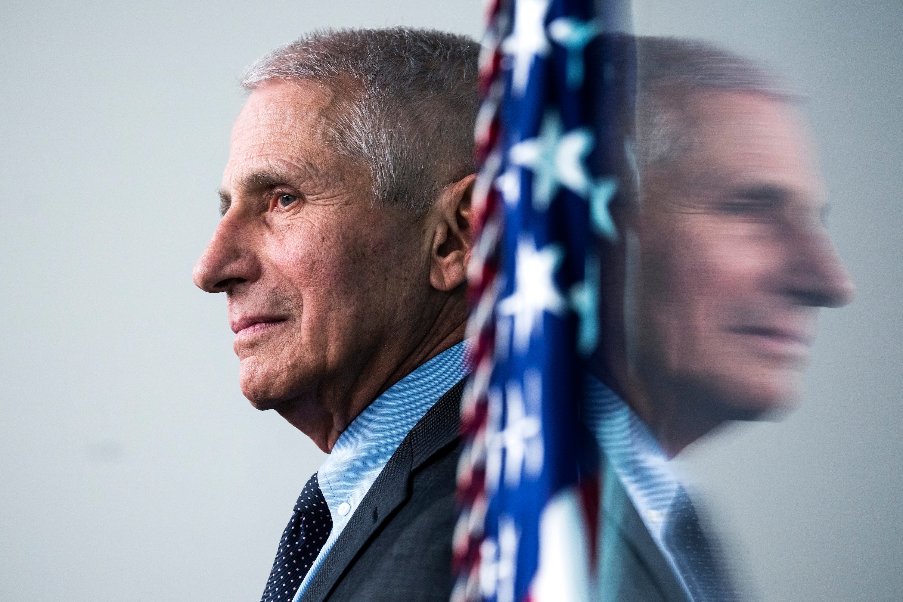 Anthony Fauci's side profile and his reflection, next to an American flag.