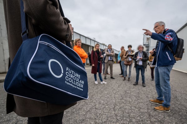 A group of people stand in a semicircle as one gestures toward something off-camera. One of them wears a tote bag emblazoned with a logo for CERN’s Future Circular Collider.