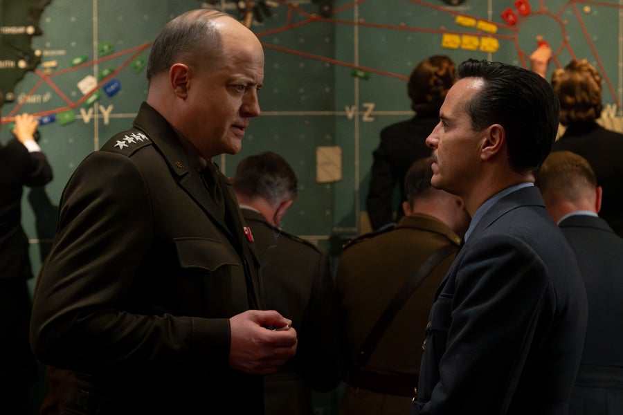 A bald man in an World War II-era U.S. army uniform speaks with another man in a blue British Royal Air Force uniform in front of a war battle map with other people behind them