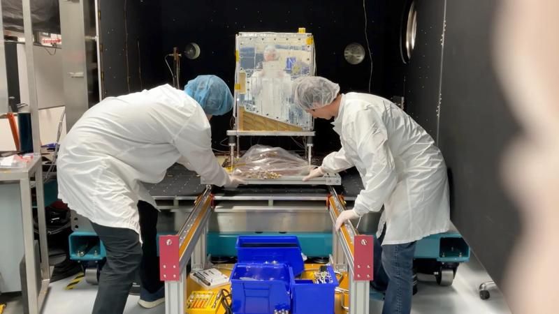 Two scientists place a satellite prototype inside a device.