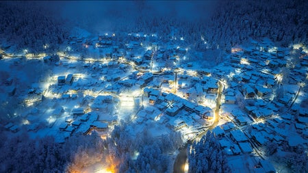 Overhead view of several houses with roofs covered in snow clustered around well-lit streets at night.