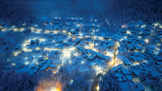 Overhead view of several houses with roofs covered in snow clustered around well-lit streets at night.