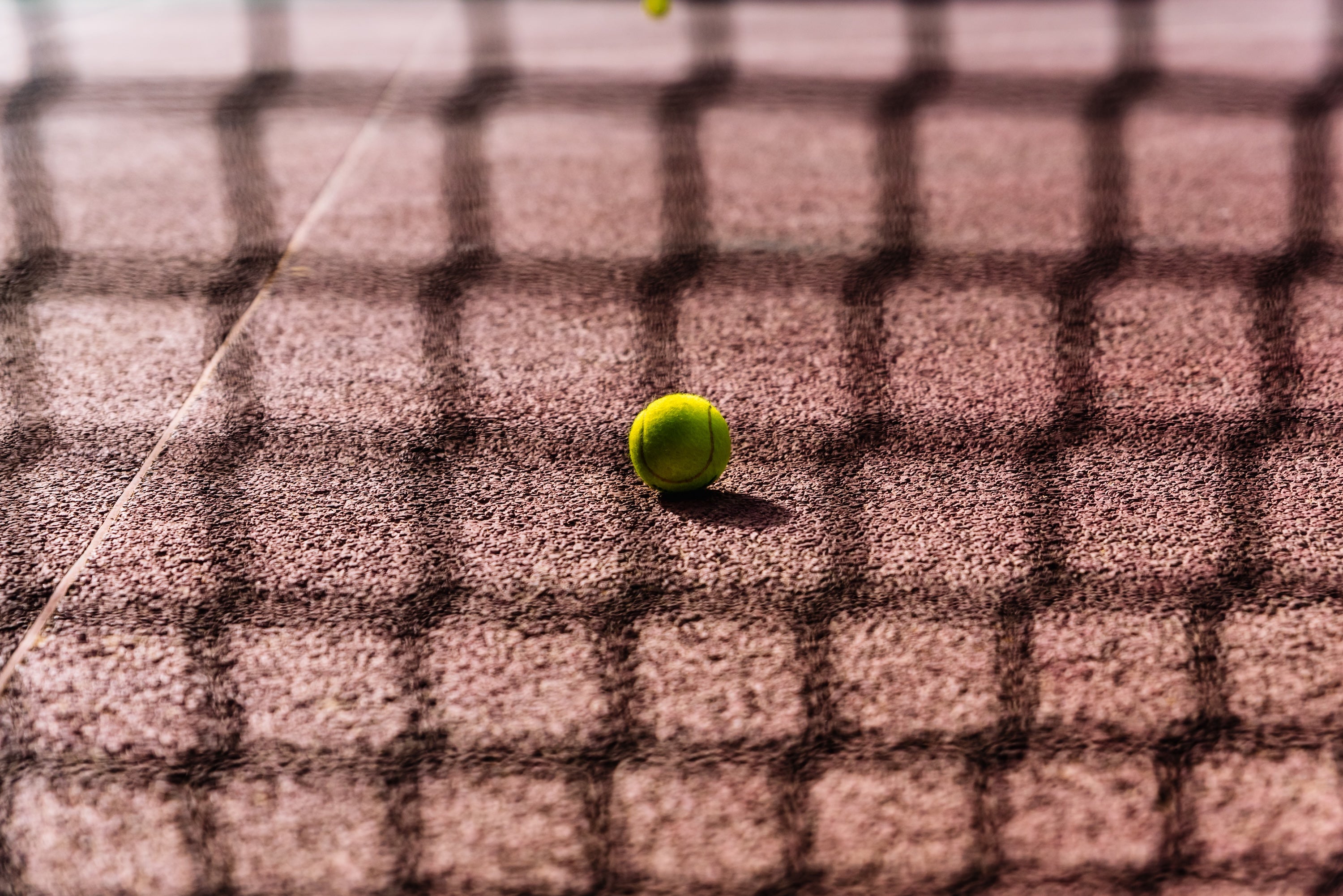 A tennis ball sits on a clay court with the shadow of the net overlapping it.