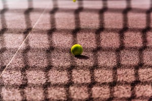 A tennis ball sits on a clay court with the shadow of the net overlapping it.