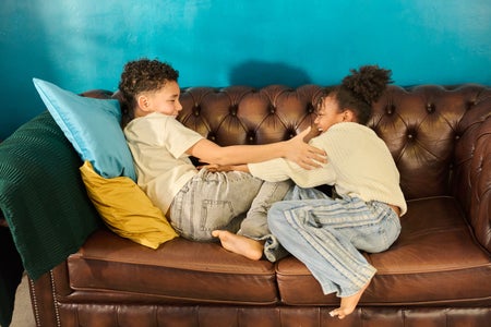 Two children playfully pushing and laughing together on a sofa in a bright home interior