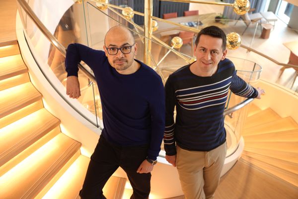 Two men face the camera from different levels of a sweeping white spiral staircase.