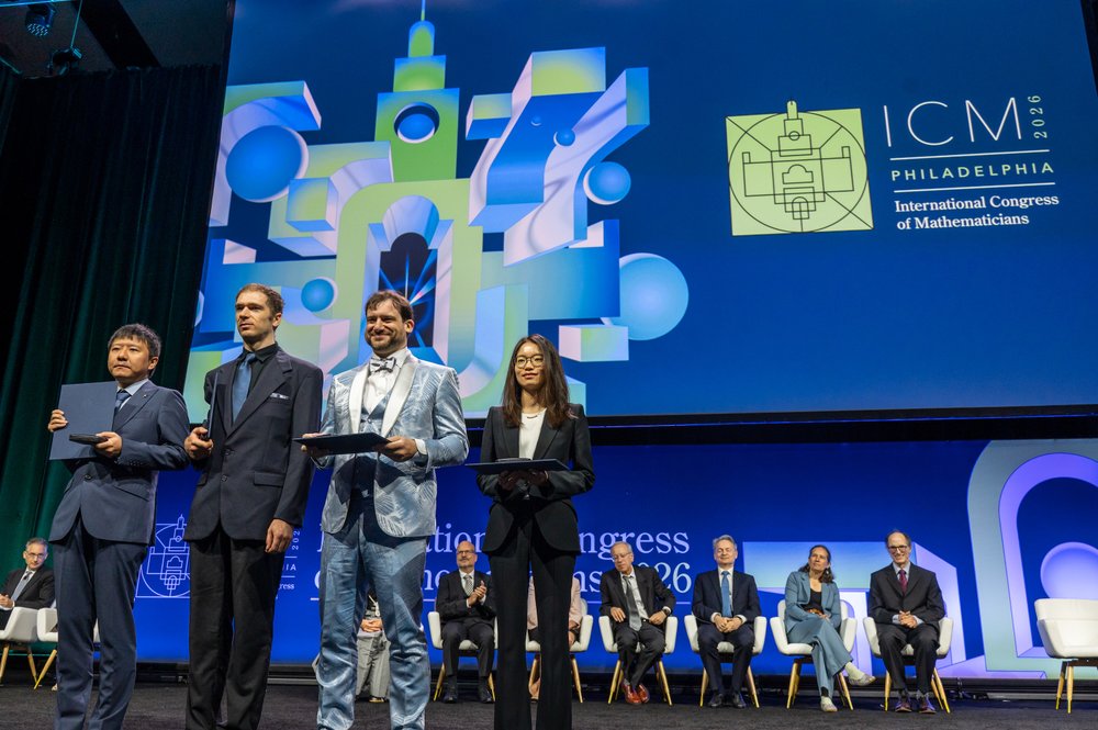 Four young mathematicians stand on a stage in a line holding folders, with the ICM 2026 logo behind them.
