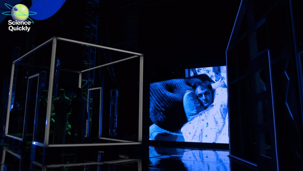 An image of a stage where people are standing in a box and image of man in a bed is being projected