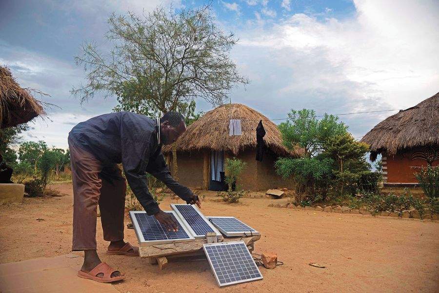 A man bends over to adjust a set of small solar panels propped on logs in the middle of a group of thatched-roof huts. Image provided by ISAAC KASAMANI/Stringer/Getty Images.