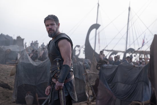 A man standing in Classical Greek armor with a ship behind him
