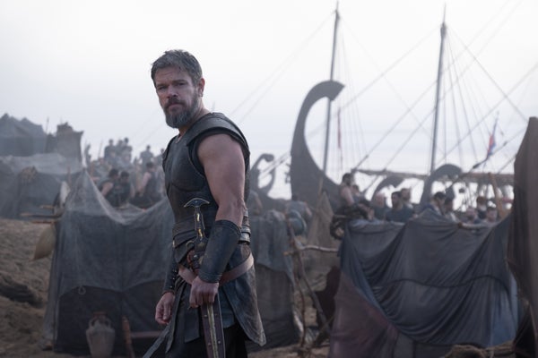A man standing in Classical Greek armor with a ship behind him