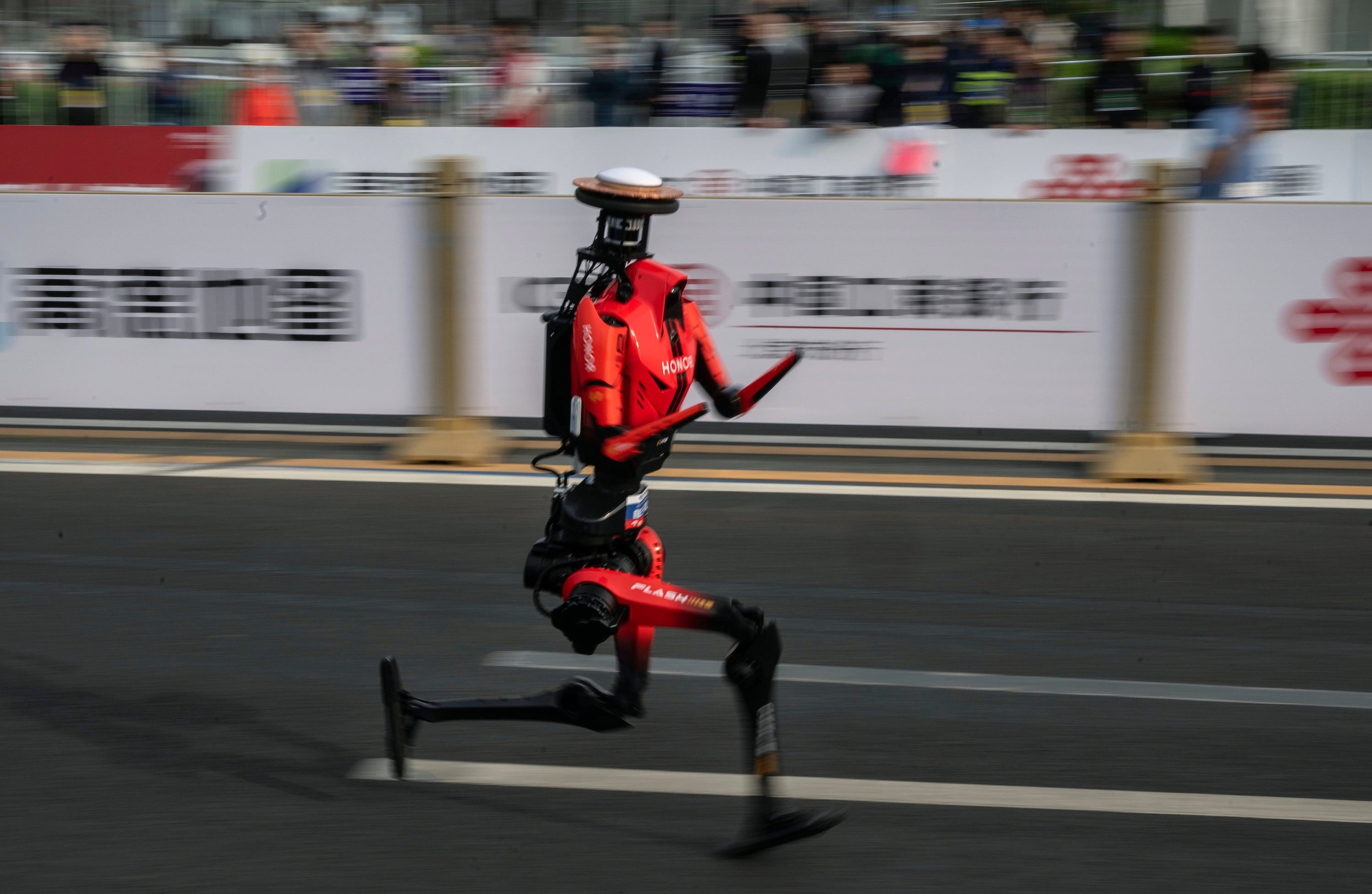 Red humanoid robot runs down a road racecourse, with the background crowd and barriers blurred by motion.