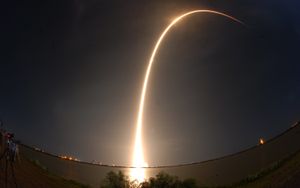 A rocket soars into space, leaving an arc of light in the dark sky.