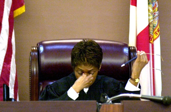 A judge sits at the bench between flags, looking down with one hand to her face and eyeglasses raised in the other.
