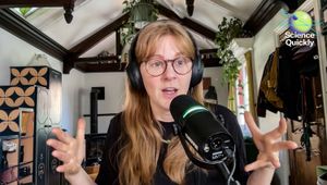 A woman in glasses speaking into a microphone with hands raised